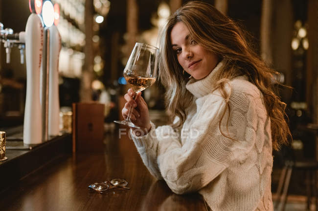 Atractiva joven en suéter de punto de moda disfrutando del vino blanco mientras se sienta en el mostrador en un elegante pub - foto de stock