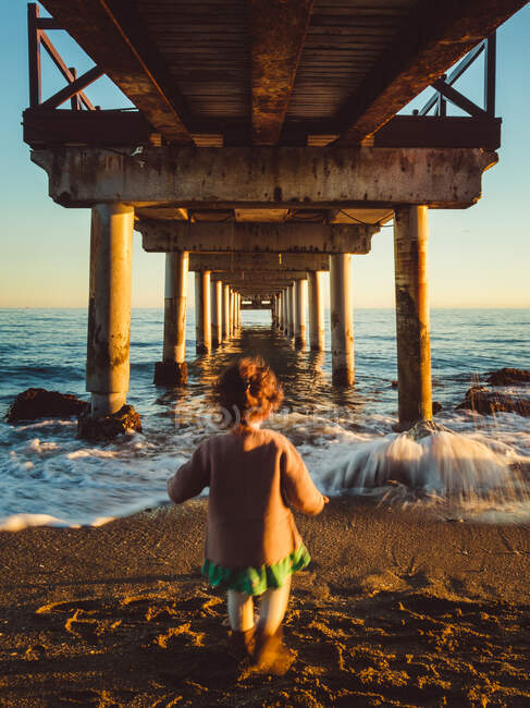 Дитина позаду ходить під пірсом на пляжі на заході сонця — стокове фото