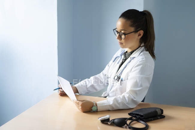 Молода жінка-лікар в уніформі дивиться на папір, сидячи за столом з тонометром в кімнаті — стокове фото