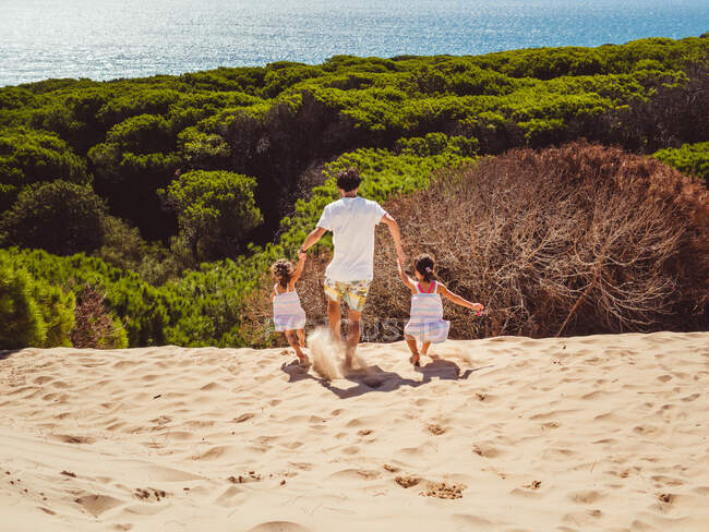 Обратный вид анонимного отца с двумя дочерьми, гуляющими по песчаной дюне в яркий летний день — стоковое фото