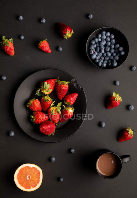 Various fresh berries with grapefruit and mug of aromatic hot beverage for breakfast on black background — Stock Photo