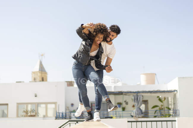 Multiracial hombre y mujer riendo y equilibrando en la pared mientras se divierten en la calle de la ciudad durante la fecha - foto de stock