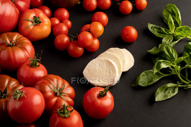 Спелые красные помидоры и листья базилика для салата на черном фоне рядом с ломтиками свежего сыра моцарелла — стоковое фото