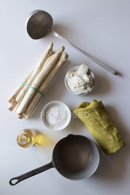 Cazo vacío y servilleta verde sobre mesa blanca cerca de manojos de espárragos frescos y crema con aceite y sal para cocinar sopa - foto de stock