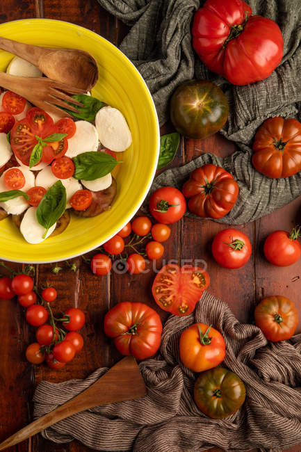 Frische reife Tomaten auf hölzerner Tischplatte in der Nähe einer Schüssel mit leckerem Caprese-Salat — Stockfoto