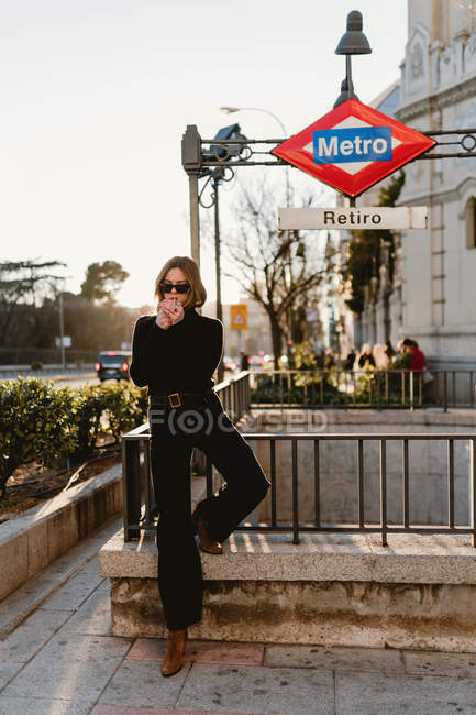 Atractiva hembra en traje de moda iluminación cigarrillo, mientras que de pie cerca de la estación de metro en la calle de la ciudad y fumar - foto de stock
