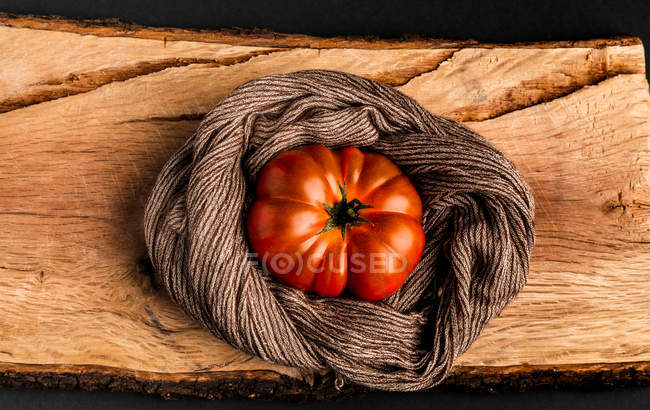 Fresh ripe tomato and fabric napkin on piece of wood on black background — Stock Photo