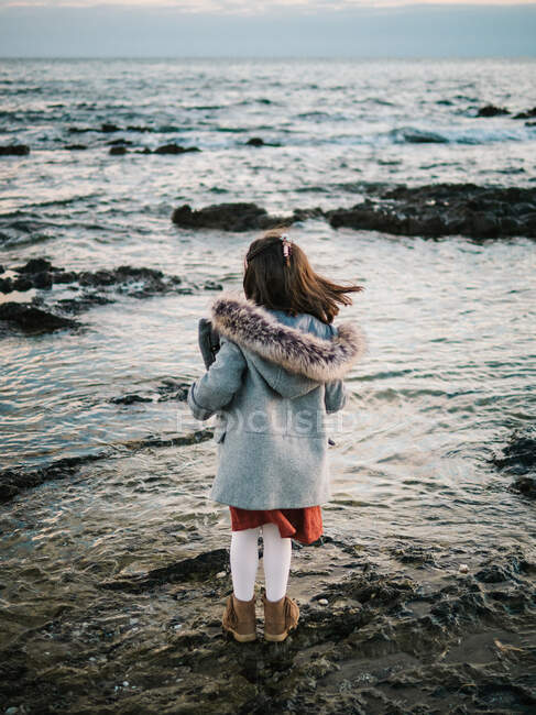 Anonima bambina da dietro in piedi vicino alla spiaggia sugli scogli guardando il mare in una giornata invernale — Foto stock
