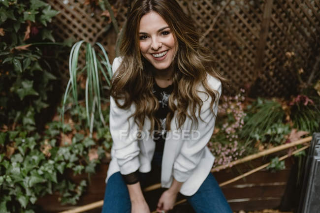 Lächelnde junge Frau in stylischem Outfit blickt in die Kamera, während sie auf einem hölzernen Pflanzkübel im wunderschönen Garten im Hinterhof sitzt — Stockfoto