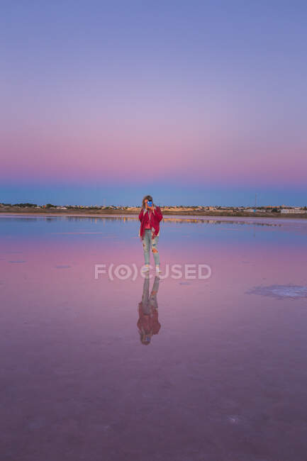 Femme prenant des photos avec caméra dans un ciel bleu rose sur le bord de mer calme vide — Photo de stock