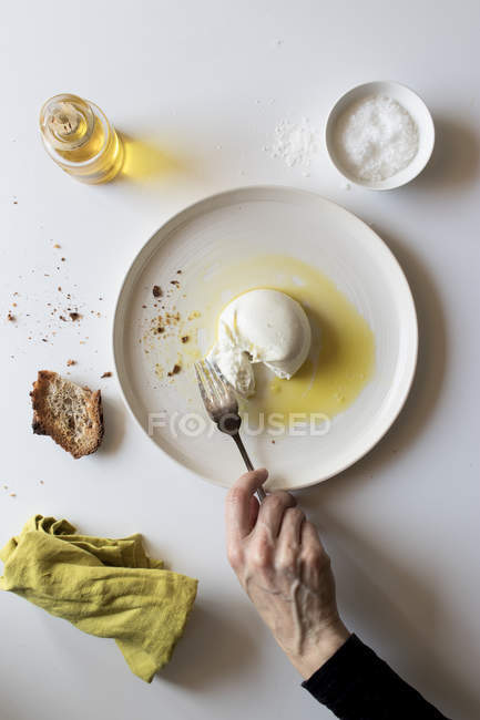 Hand eines anonymen Seniors mit Gabel, um ein Stück leckere frische Burrata von einem Teller in der Nähe von Brot und Öl vor weißem Hintergrund zu nehmen — Stockfoto