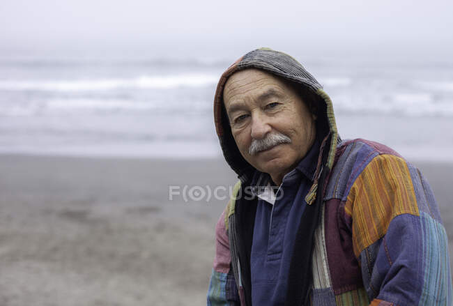 Vista lateral do homem sênior de casaco em pé na praia vazia remota da costa oceânica olhando para a câmera — Fotografia de Stock