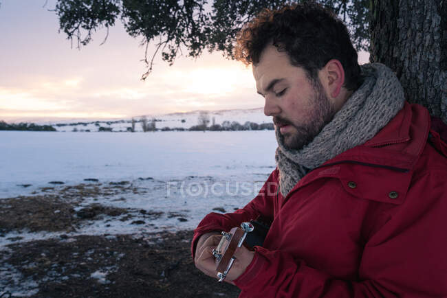 Vue latérale d'un homme d'âge moyen en vêtements chauds jouant de la guitare sous un arbre sur fond de paysage enneigé — Photo de stock