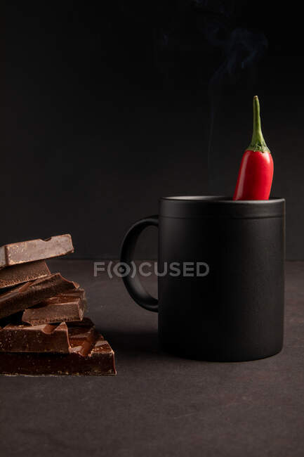 Trozos de delicioso chocolate colocados cerca de la taza negra de bebida caliente humeante con chile sobre fondo oscuro - foto de stock