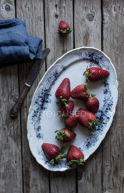 Teller mit leckeren reifen Erdbeeren auf hölzerner Tischplatte neben blauer Serviette und Metallmesser — Stockfoto