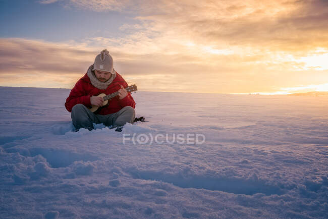 Человек в теплой одежде сидит на просторном снежном поле и играет на гитаре на фоне яркого закатного неба — стоковое фото