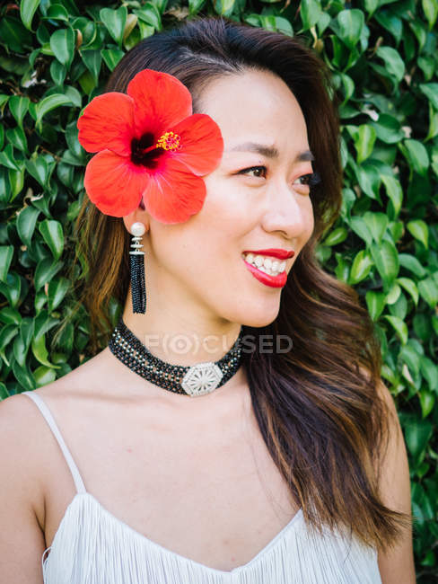 Porträt einer jungen eleganten Chinesin vor Blätterhintergrund mit roter Blume im Kopf — Stockfoto