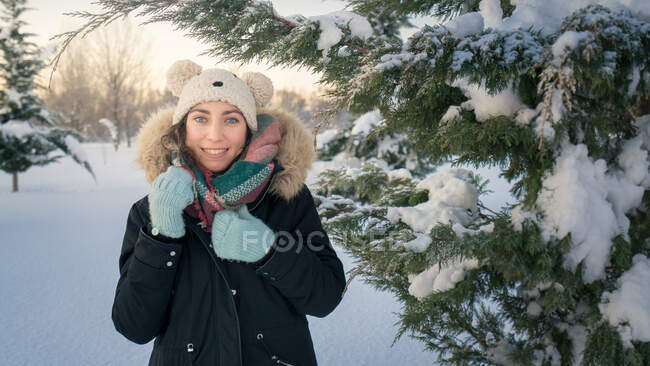 Jovem fêmea atraente em roupas quentes com pele alegre rindo ao lado coberto de neve conífera árvore — Fotografia de Stock
