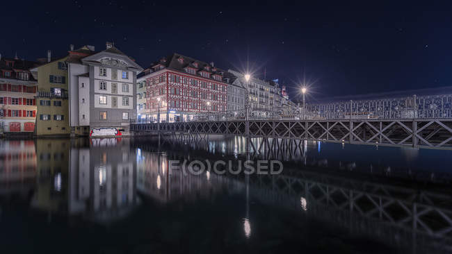 Exterior of modern buildings and bridge across calm river water illuminated in night time, Switzerland — Stock Photo