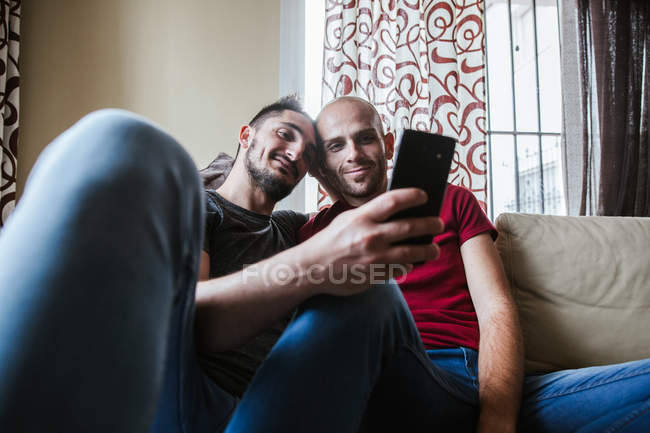 Happy gay couple relaxing on couch and sharing smartphone — Stock Photo