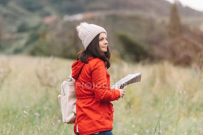 Жінка в теплому одязі тримає путівник буклетом і дивиться далеко під час прогулянки на лузі — стокове фото