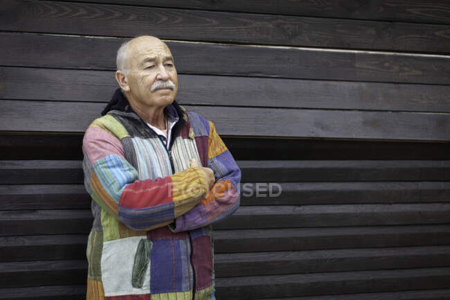 Thoughtful senior man wearing simple colorful jacket and standing with arms crossed against house wooden wall — Stock Photo