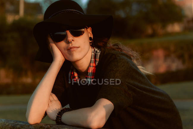 Mujer pensativa con gafas de sol de moda con sombrero negro apoyado en la mano a la luz del sol - foto de stock