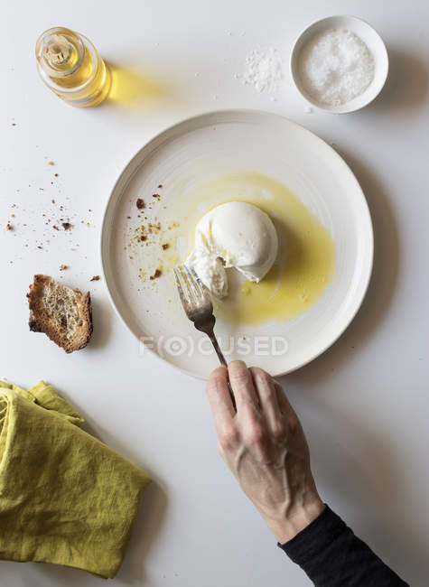 Рука анонімної людини, що тримає виделку над шматочком смачної свіжої бурата на тарілці біля хліба та олії на білому тлі — стокове фото