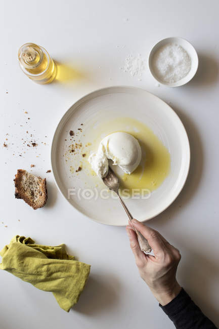 Main d'une personne anonyme tenant une fourchette sur un morceau de burrata frais délicieux sur une assiette près du pain et de l'huile sur fond blanc — Photo de stock