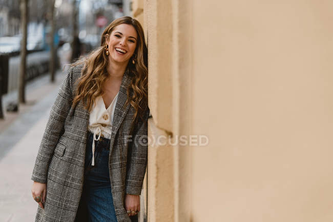 Красивая женщина в модном пальто смеется и смотрит в камеру, опираясь на стену здания на размытом фоне городской улицы — стоковое фото