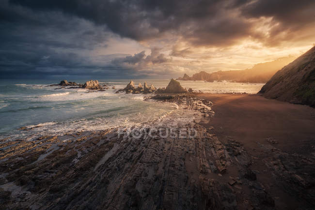 Сценический вид пустого пляжа с камнями и волнами на фоне заката с дождевыми облаками — стоковое фото