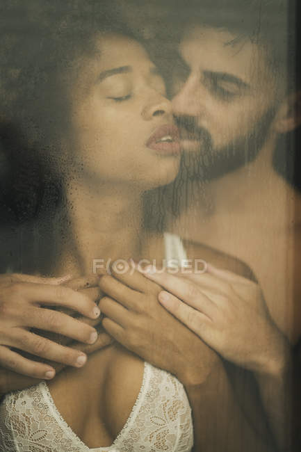 Hermoso chico hispano tocando y besando seductora mujer afroamericana en sujetador de encaje mientras está de pie detrás de una ventana húmeda - foto de stock