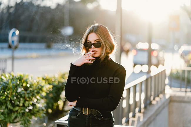 Mulher elegante fumar cigarro perto da estação de metro na rua ensolarada — Fotografia de Stock