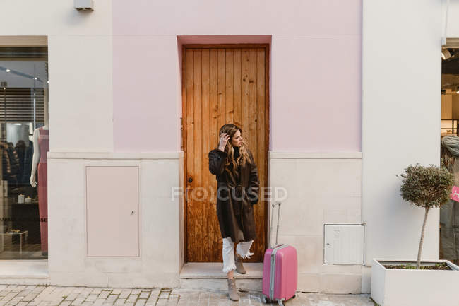 Стильна жінка з валізою, що стоїть біля коричневих дверей — стокове фото