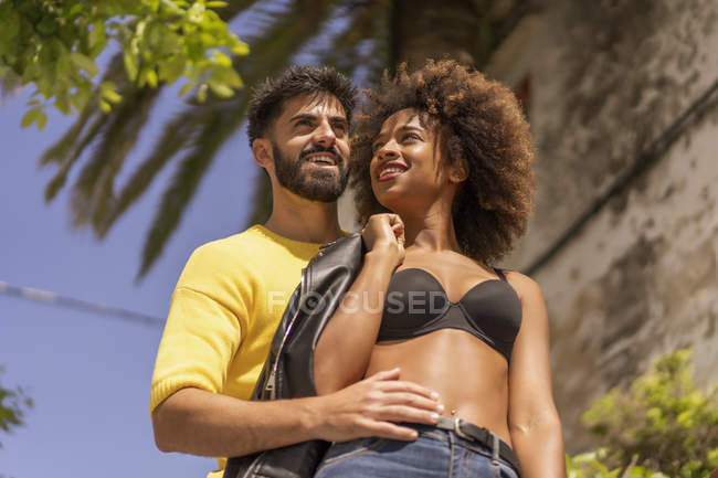 Красивый бородатый парень улыбается и флиртует с привлекательной черной женщиной в лифчике, стоя вместе на городской улице в солнечный день — стоковое фото