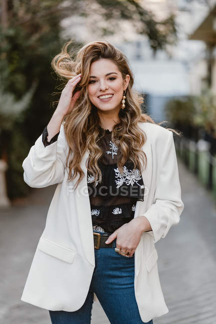Усміхнена молода жінка в модному вбранні торкається волосся, стоячи на розмитому тлі міської вулиці — стокове фото