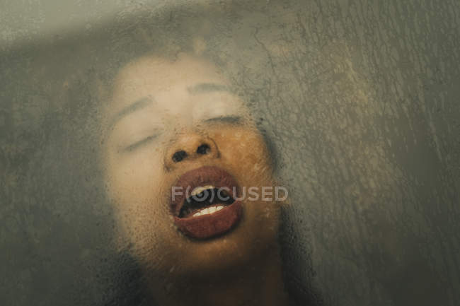 Sensual black female with closed eyes moaning and breathing heavily while having sex behind wet window — Stock Photo