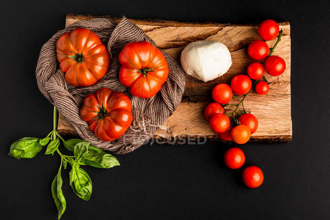 Tomates maduros frescos, mussarela e folhas de manjericão em pedaço de madeira sobre fundo preto — Fotografia de Stock