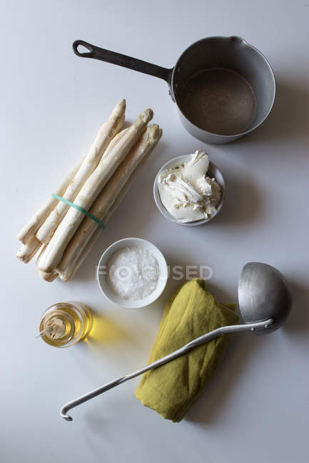Panela vazia e guardanapo verde na mesa branca perto de um monte de espargos frescos e creme com óleo e sal para cozinhar sopa — Fotografia de Stock