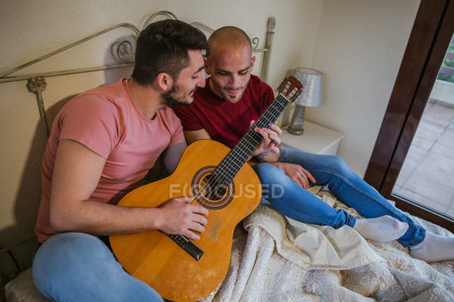 Веселая гей-пара, играющая на гитаре в спальне — стоковое фото