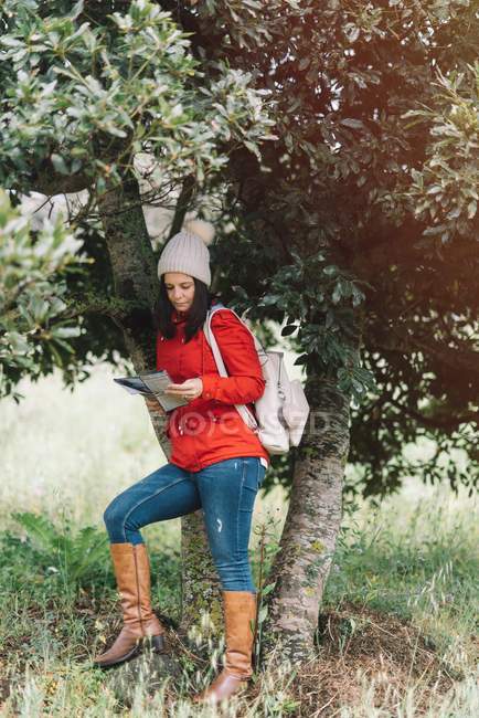 Красива жінка в теплому одязі читає путівник по дереву в природі — стокове фото