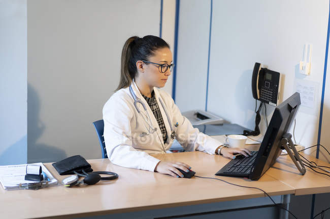 Молодая женщина врач в форме просмотра на компьютере и сидя за столом с тонометром в комнате — стоковое фото