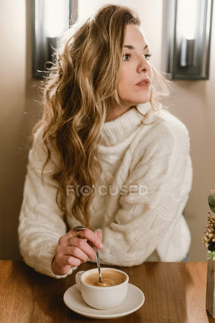 Чуттєва жінка в стильному в'язаному светрі, дивлячись в сторону і перемішуючи ароматну каву, сидячи за столом кафе — стокове фото