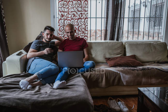 Feliz gay casal relaxante no sofá e usando smartphone e laptop — Fotografia de Stock