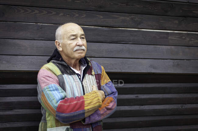 Thoughtful senior man wearing simple colorful jacket and standing with arms crossed against house wooden wall — Stock Photo