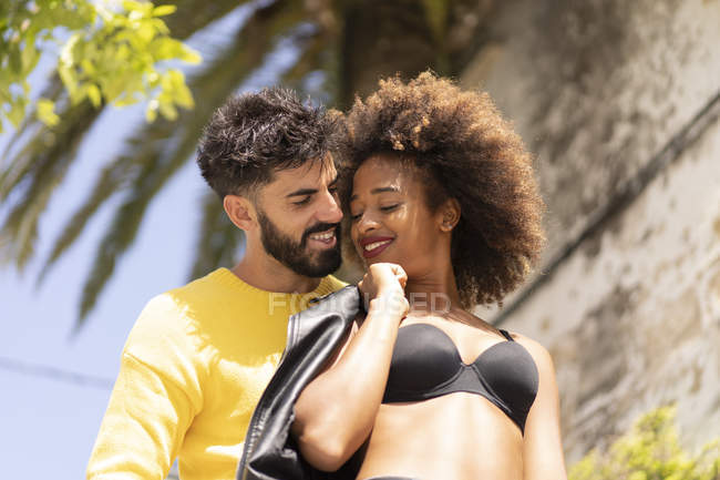Красивий бородатий хлопець посміхається і фліртує з привабливою чорною жінкою в бюстгальтері, стоячи на міській вулиці разом у сонячний день — стокове фото