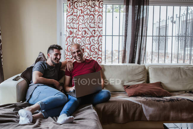 Весела гей пара використовує ноутбук, розслабляючись на дивані — стокове фото