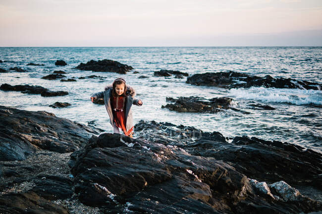 Cute little girl in winter clothes standing on a rocky beach — Stock Photo