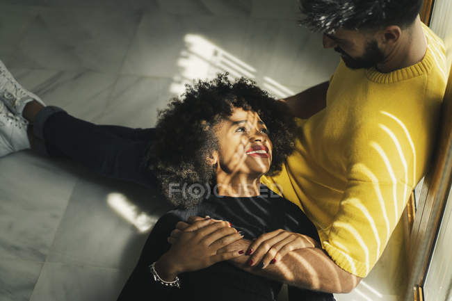 Pretty smiling African American woman laughing while lying on knees of bearded man on floor — Stock Photo
