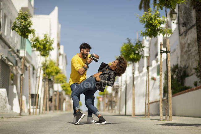 Beau barbu gars en utilisant un appareil photo pour prendre une photo de la femme criant et se penchant en arrière sur la rue de la ville — Photo de stock